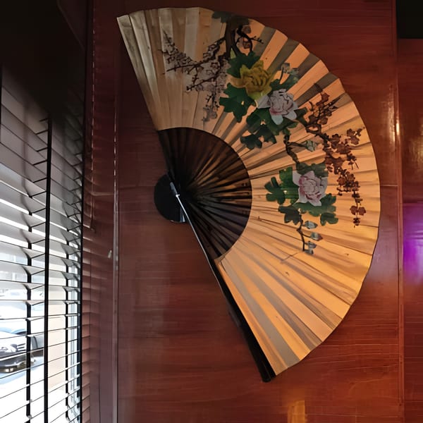 A large, decorative Asian fan with floral paintings hangs on a wooden wall next to window blinds.