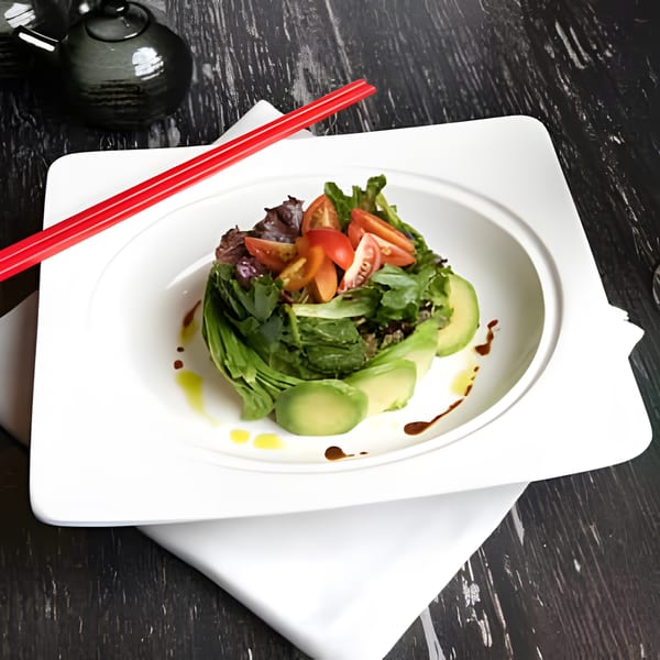 Fresh green salad with avocado slices, cherry tomatoes, and dressing, served with red chopsticks.