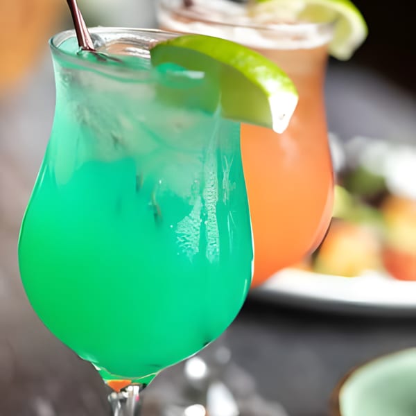 Vibrant green cocktail with a lime wedge and straw, with an orange drink blurred in the background.